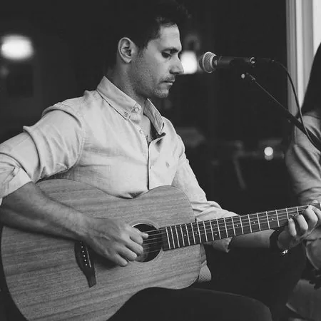 Stelios Antoniou singing into a microphone while playing acoustic guitar at night
